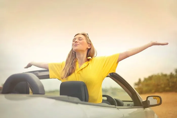 身穿黄色衬衫的年轻女子坐在敞篷银色轿车上，张开双臂享受天气