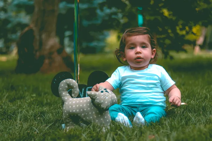 一个蹒跚学步的孩子穿着青色T恤和青色裤子，旁边是灰色的猫毛绒玩具