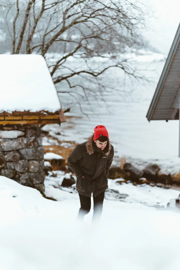 一名男子穿着棕色皮毛装饰连帽夹克，戴着红色针织帽，白天在雪地上行走