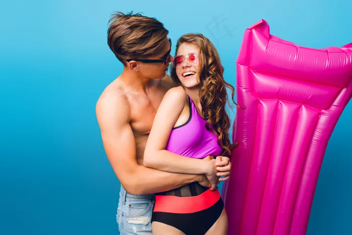 在工作室里，一对年轻的帅哥在蓝色背景下拥抱着穿着泳装的漂亮女孩。他们戴着太阳镜，女孩对男孩微笑。