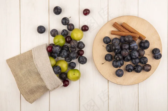 俯视图小酸蓝黑色的浆果在木制的厨房板上，肉桂棒上有绿色的樱桃李子和红色的樱桃从一个麻袋里掉出来，背景是白色的木质