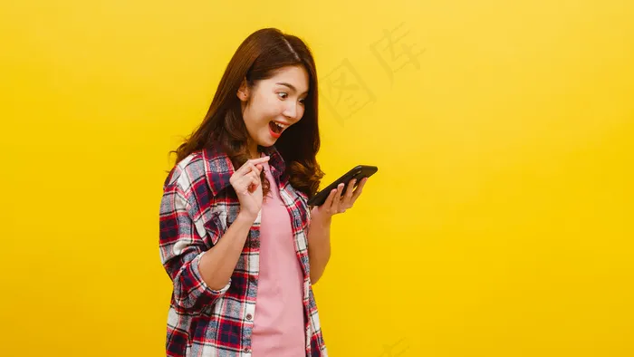照片中的亚洲女性惊讶地使用手机，表情积极，穿着休闲服，看着黄色墙上的相机。快乐可爱快乐的女人为成功而高兴。