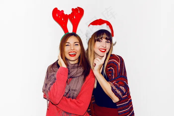 新年晚会。两个漂亮的女孩戴着滑稽的化装圣诞老人帽送上吻。最好的朋友摆姿势的室内库存图片。隔离。