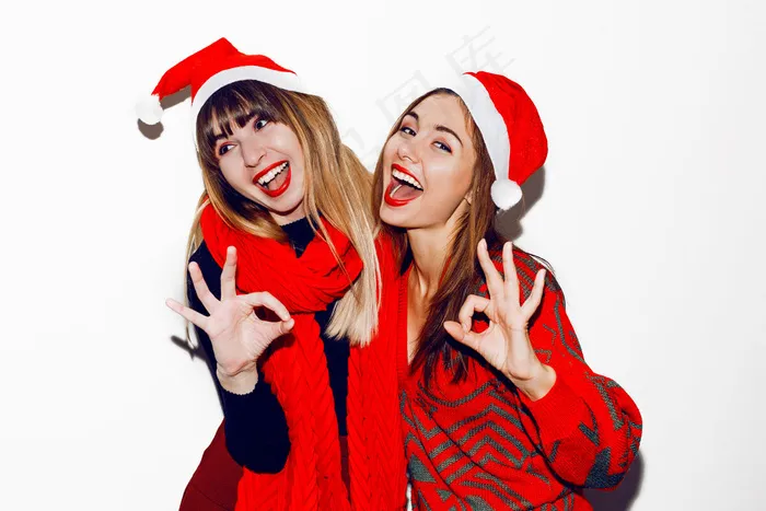 疯狂的新年派对气氛。两个醉醺醺的笑着的女人玩得很开心，还戴着可爱的化妆帽摆姿势。红毛衣和围巾。用手表示可以。