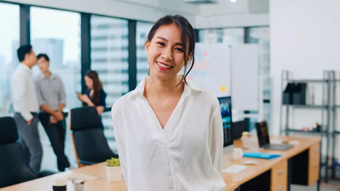 在现代办公场所，一位成功的漂亮的女商务人士在照相机前微笑。站在当代会议室的年轻亚洲女士。