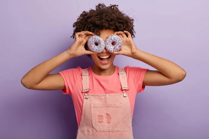 美丽有趣的非裔美国女人眼睛上戴着甜美的紫色甜甜圈，在室内享受美味的甜点，穿着粉色的衣服，在紫色的背景下与世隔绝。节食，垃圾食品，减肥概念