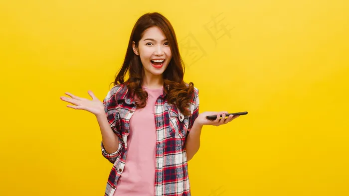 照片中的亚洲女性惊讶地使用手机，表情积极，穿着休闲服，看着黄色墙上的相机。快乐可爱快乐的女人为成功而高兴。