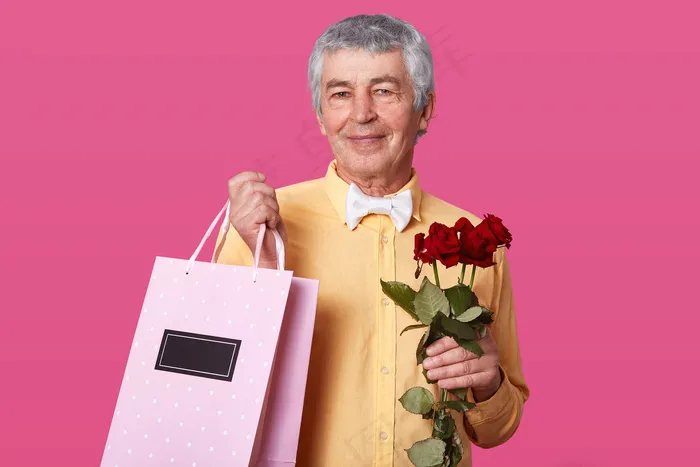 照片中，一张漂亮的成熟男子，表情愉快，身穿黄色衬衫，搭配白色蝴蝶结，拎着粉色的包，带着礼物和玫瑰，想祝贺妻子结婚周年。